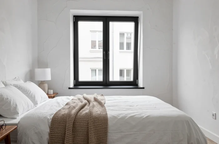 Chambre de style scandinave avec fenêtre oscillo-battante à cadre aluminium noir mat, literie blanche et plaid en laine épaisse couleur avoine